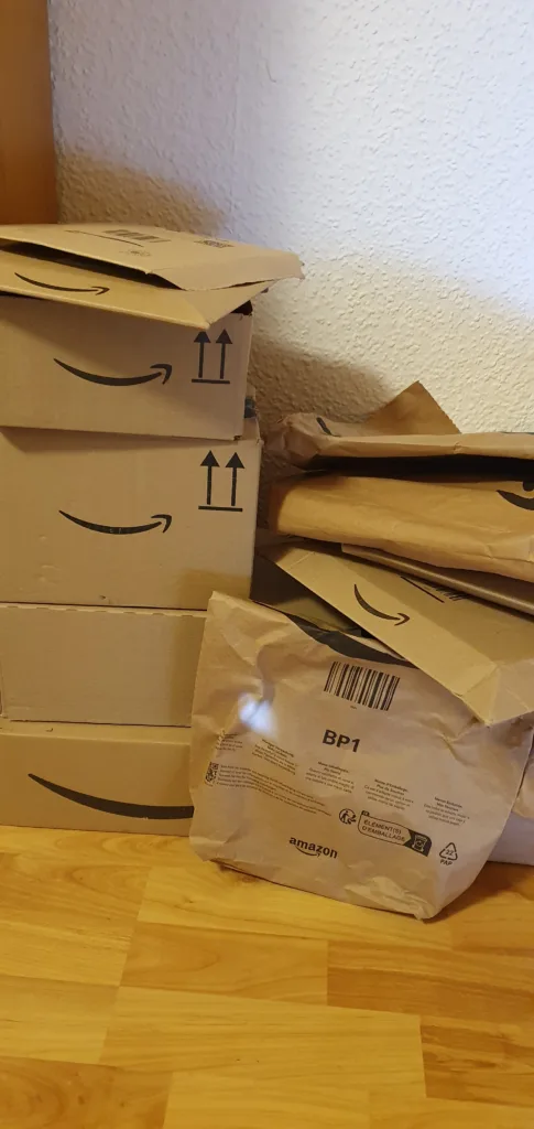 Amazon-Kartons in einer Zimmerecke, davor eine gepolsterte Versandtasche auf Parkettboden